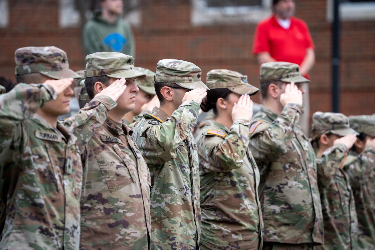 Six soldiers in camouflage are lined up with their right hands raised in salutes.