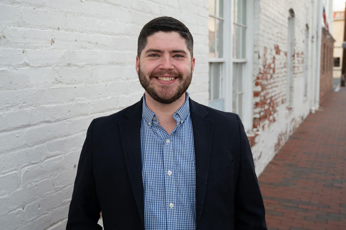 This is a portrait of Chris Scroggins who is standing outside along a white brick building with windows in the background. Chris is wearing a blue plaid shirt and dark suit jacket and has a beard, mustached and short dark hair.