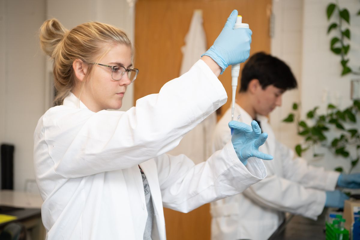 Photograph of two young students, one female and one male, working in a lab wearing white lab coats. The female has blonde hair and is wearing glasses. She is using a syringe to add something to a tiny test tube. 