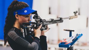 A person stands on the left side of this photo while holding a competition rifle. The person has long dark hair and is wearing a blue visor and black top.
