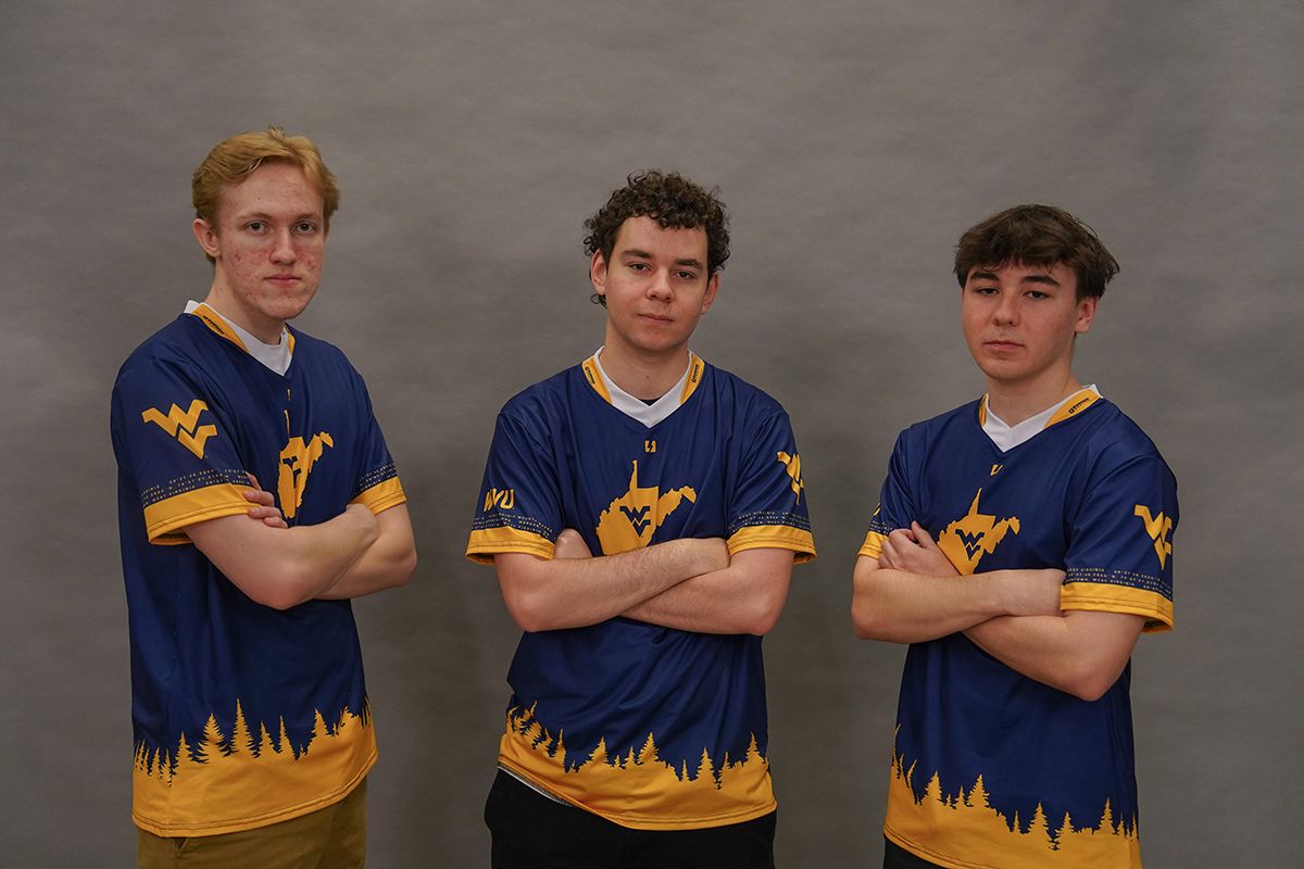 Three members of the WVU Esports Rocket League team wear gold and blue shirts while standing in front of a gray back drop.