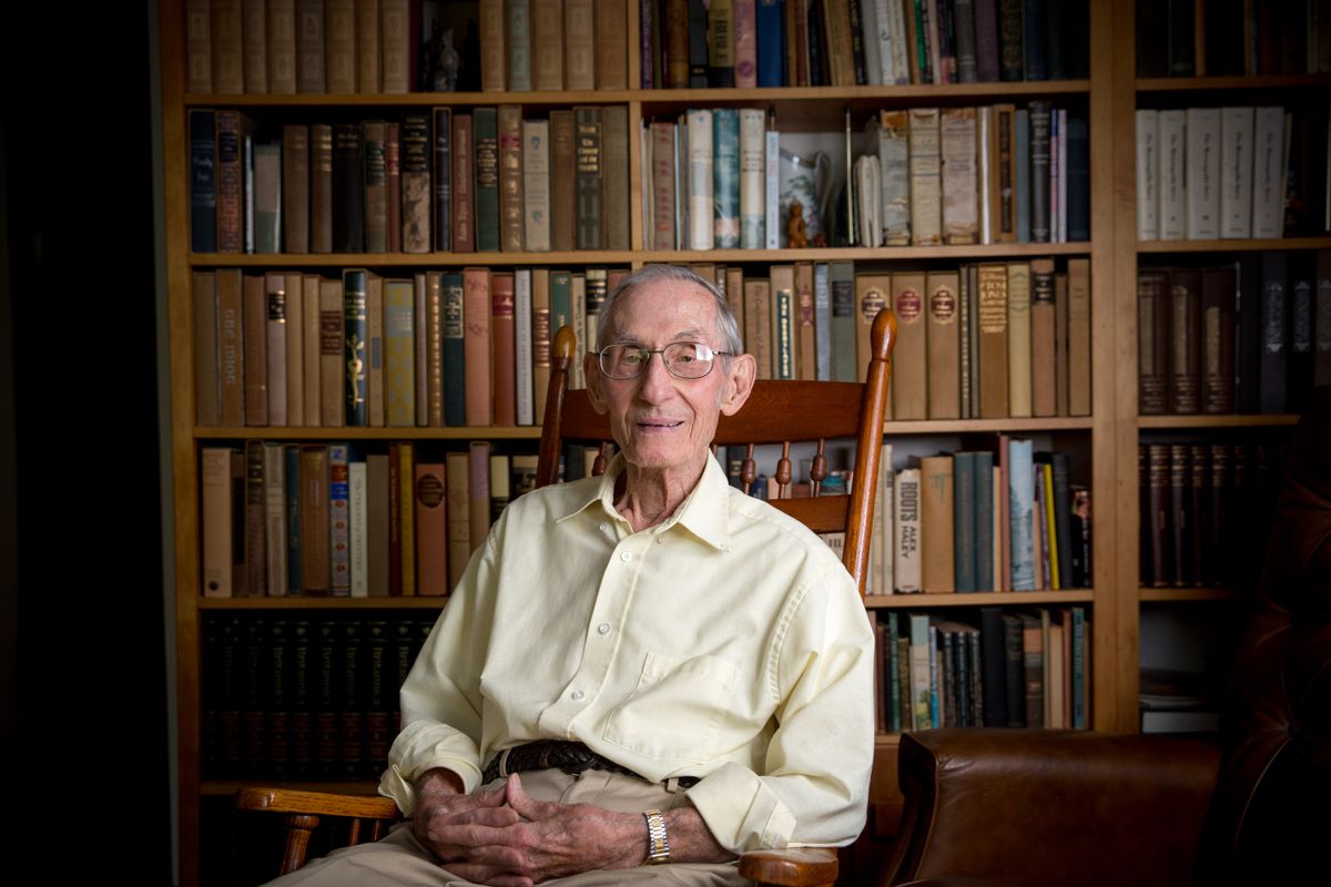 Photo of an older man in long-sleeved off white shirt sitting in a chair in front of shelves of books
