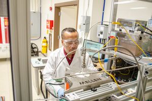 A WVU researcher works on scientific equipment wearing a white lab coat, blue safety gloves and safety glasses. 