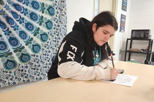 A student at Preston High School sits at a desk holding a pencil in their right hand and writing on a white sheet of paper. The students long dark hair and is wearing a black sweatshirt with long white sleeves. A dark blue, teal tapestry is on the wall.