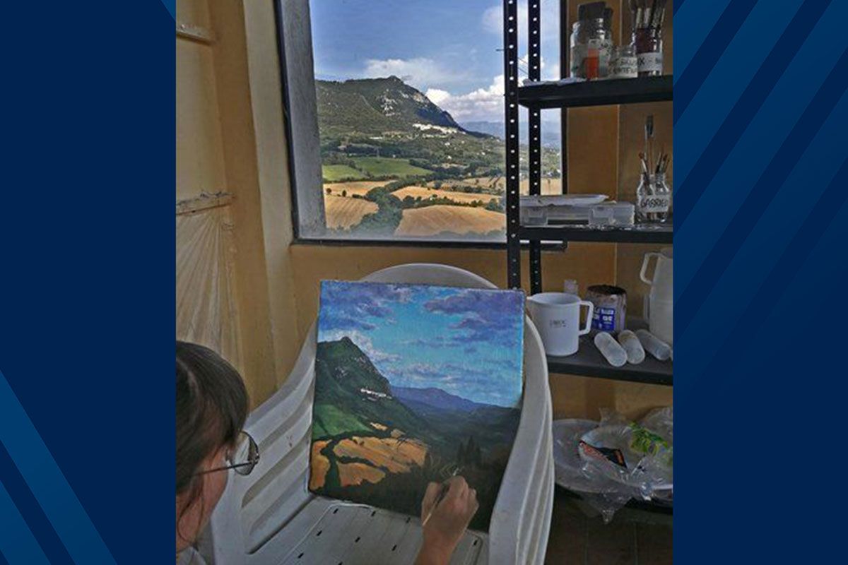 A student looking out the window painting a picture of the landscape.