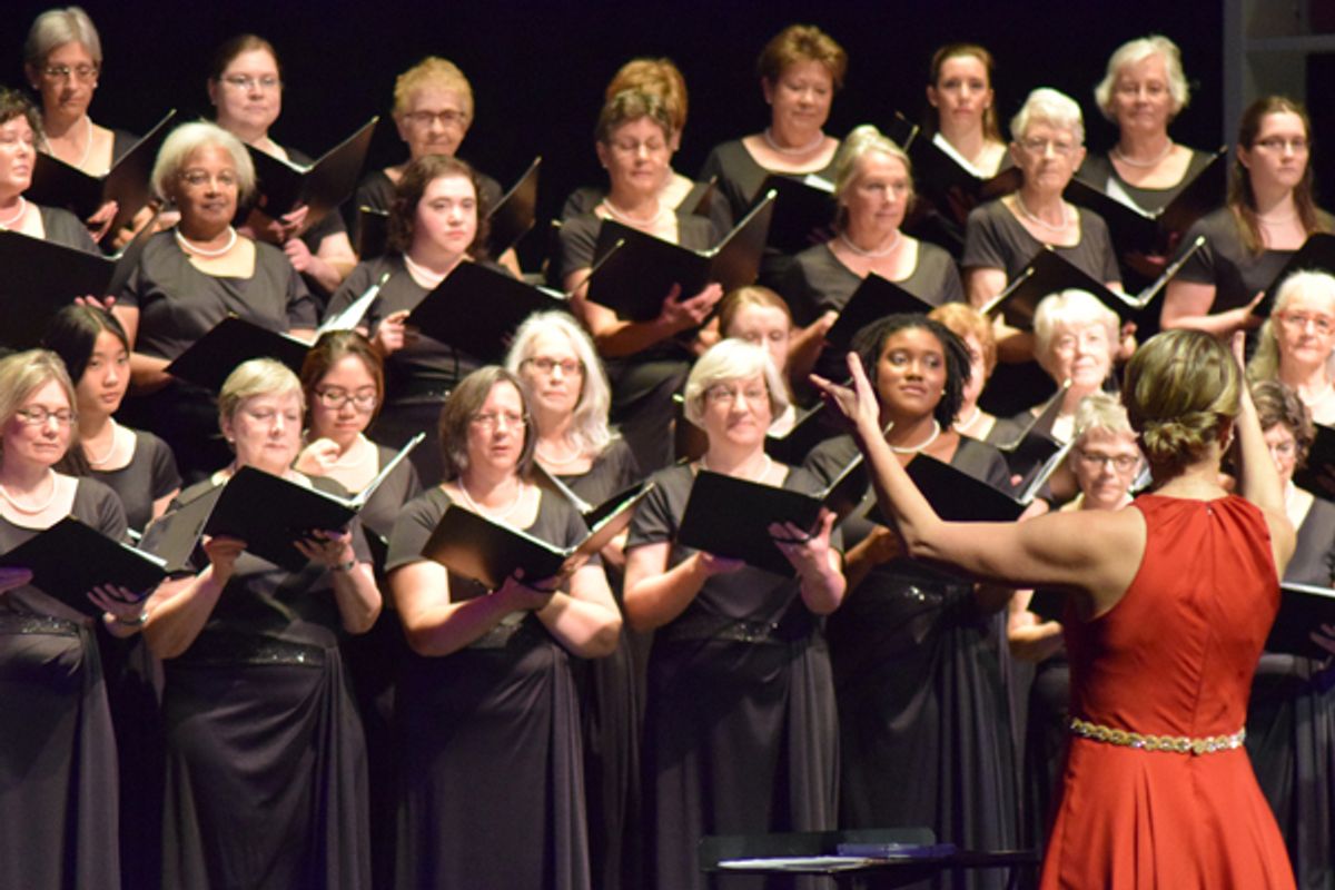 Photo of a woman in a sleeveless red dress conducts a choir of women in black dresses