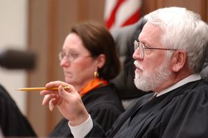Larry Starcher is shown from the left side while presiding from the bench. He is wearing a dark robe, has gray hair and glasses and is pointing with a pencil in his right hand.