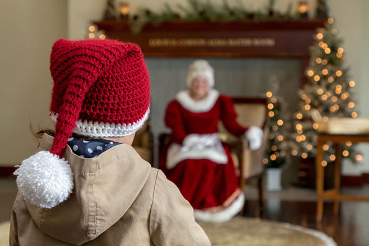 A young boy approaching Mrs. Clause seated on a chair.